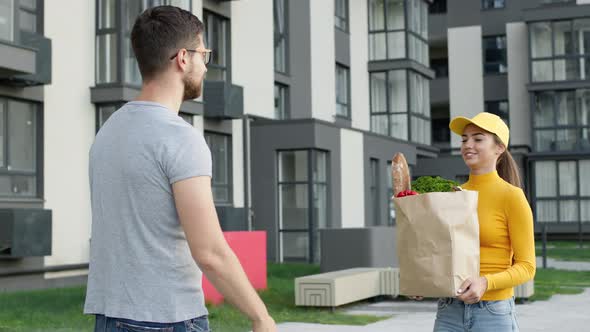 Goods Delivery. Girl Delivery Service Worker Gives Guy Package Of Groceries.