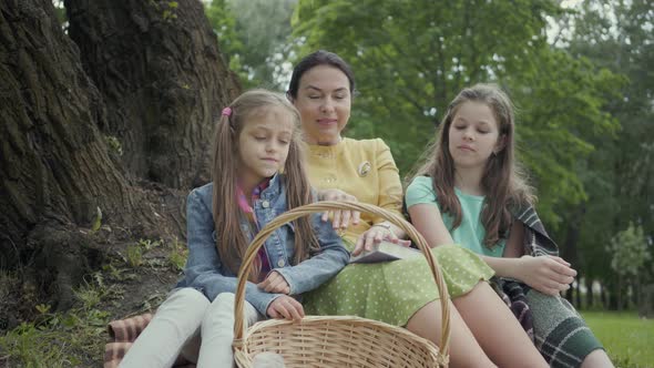 Mature Woman Sitting on the Grass Under the Tree in the Park with Two Cute Granddaughters