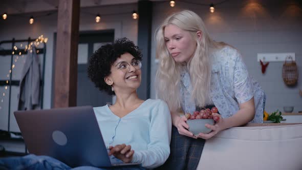 Young Girls Looks at the Laptop and Laugh Girl Leans on the Sofa Holding Food