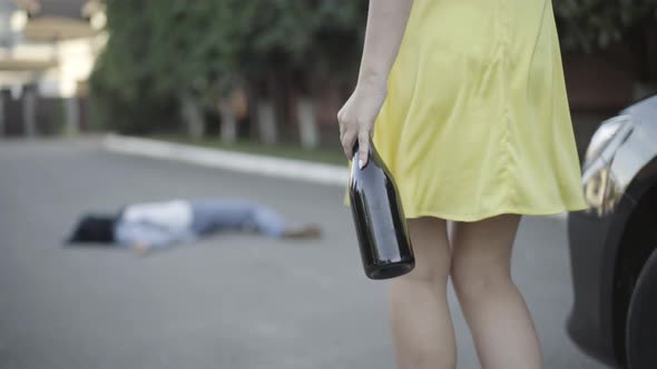 Camera Follows Unrecognizable Drunk Caucasian Woman with Wine Bottle Walking To Corpse