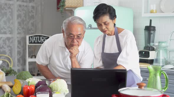 70s Elderly Senior Asian Couple Watching Online Tutorial on Laptop While Cooking in Kitchen at Home