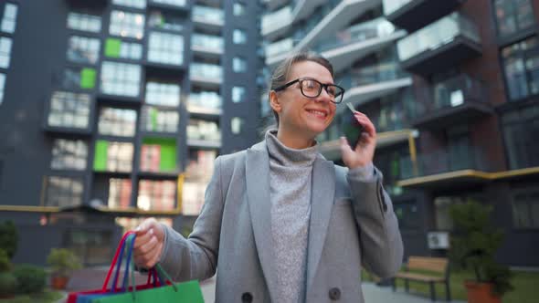 Smiling Woman Walking on a Business District with Shopping Bags and Talking on the Smartphone