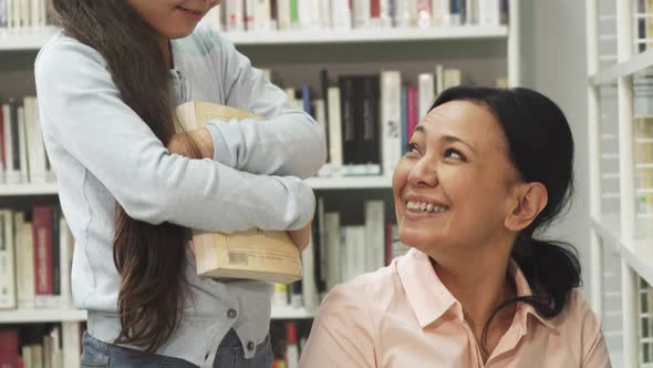 Pretty Little Girl Smiling While Her Mom Reading a Book at the Library