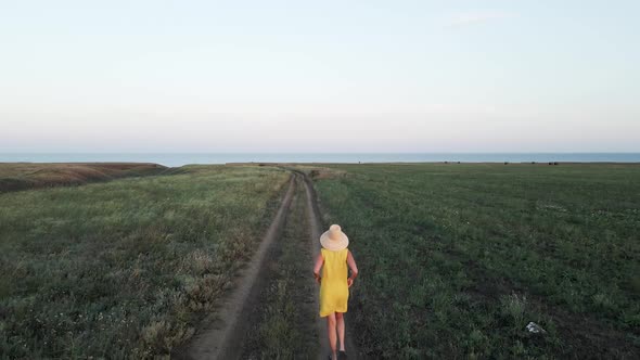 Woman in a Yellow Dress and a Straw Hat Walks Through a Field and a Dirt Road