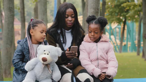 African American Happy Family Sitting on Bench in Park Mom with Daughters Look at Phone Little Girl