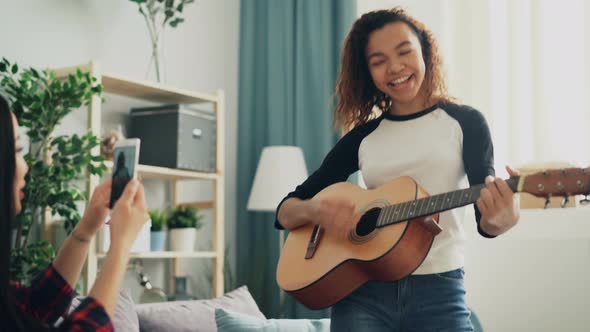 Adorable Young Woman Amateur Guitarist Is Playing the Guitar and Singing While Her Asian Friend Is