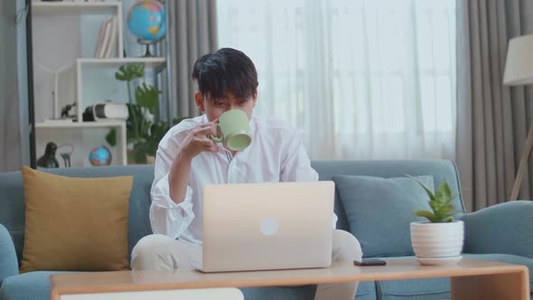 Happy Asian Man Drinking Coffee While Using Laptop Computer In The Living Room