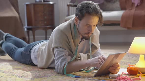 Young Creative Man Searching Inspiration Online Surfing Internet on Tablet Lying on Floor in Atelier