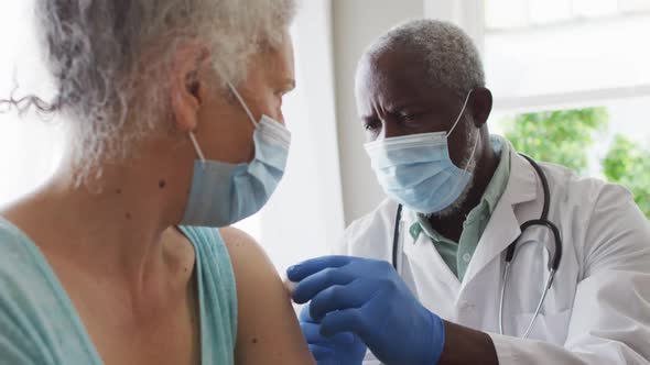 Male african american doctor wearing face mask injecting covid-19 vaccine into senior female patient