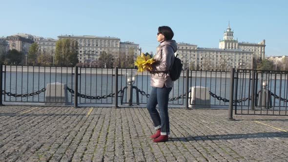 A Middleaged Woman Brunette with a Bouquet of Yellow Leaves Walks Along the City Embankment