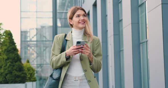 Cheerful Young Caucasian Woman Walking in the Street to the Office with Cellphone in Hand