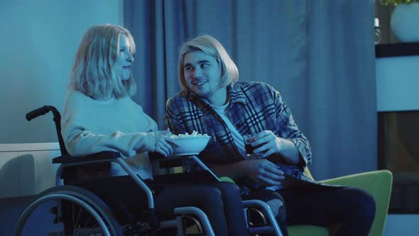 Young Man and Woman in a Wheelchair Watching TV  Eating Popcorn and Drinking Soda