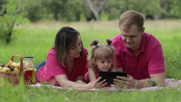 Family Weekend Picnic. Daughter Child Girl with Mother and Father Play Online Games on Tablet