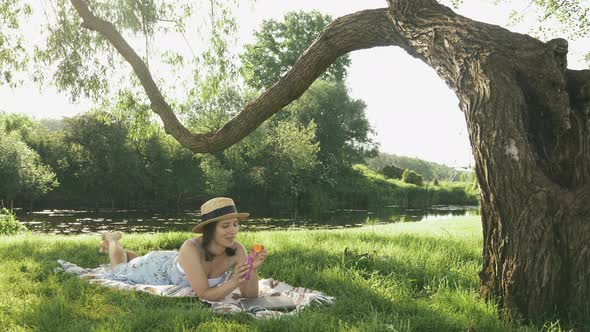 Cute girl relaxing on blanket on grass under big tree and blowing bubbles at sunny summer day