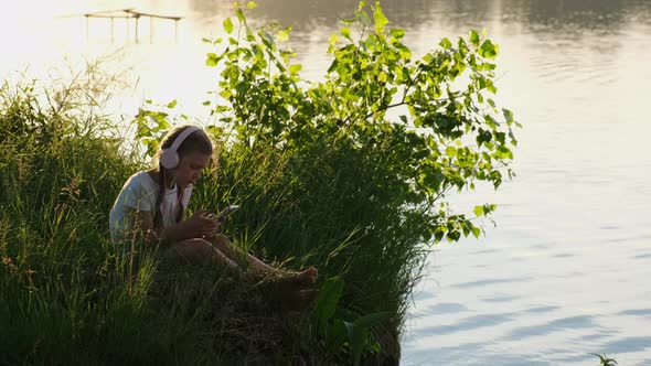 A little girl in headphones with a smartphone listening to music on the lake during the sunset.