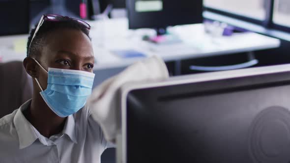 African american woman wearing face mask cleaning her computer with cloth while sitting on her desk