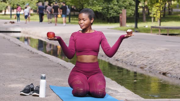 Attractive Young Adult Woman Fitness Instructor Sitting on Yoga Mat in Summer Park Holds Apple and