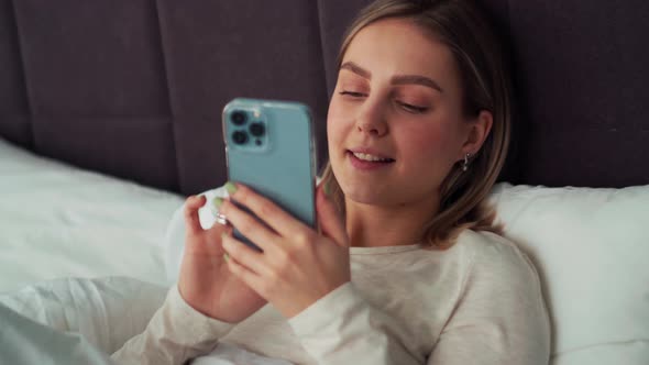 Cheerful blonde woman texting by phone in the bed