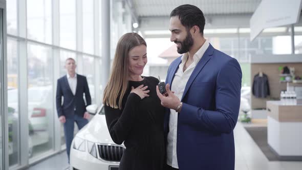 Happy Young Caucasian Woman Talking Car Keys From Middle Eastern Man in Dealership and Smiling at