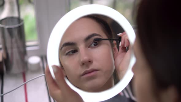 A Young Woman Looks Into a Small Illuminated Mirror and Paints Her Eyelashes with Mascara. View of