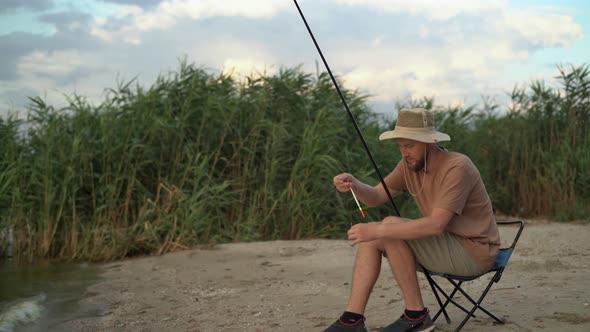 a Man Fisherman at Sunset in a Hat on the Lake Catches Fish with a Fishing Rod Against the