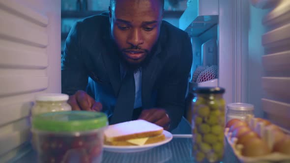 Hungry AfricanAmerican Entrepreneur Open Fridge and Eat Sandwich in Evening