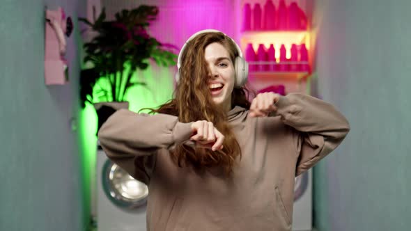 Funny Young Woman with Curly Brown Hair in Beige Headphones and Beige Sports Suit Dances Against