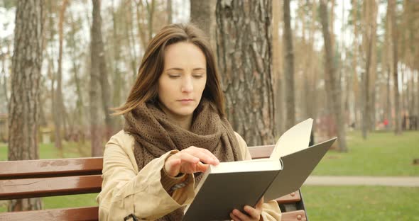 Cute focused woman turns pages of book and reading while sitting on bench in autumn cold park alone