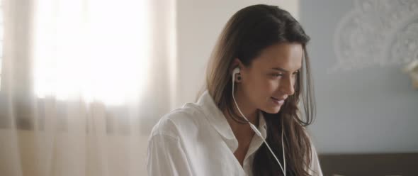 Close up of a young woman talking to someone through earphones while sitting on bed. 