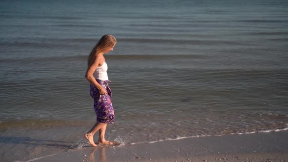 Slow motion of mature woman in sarong walking on a beach and splashing in the water at sunrise or su