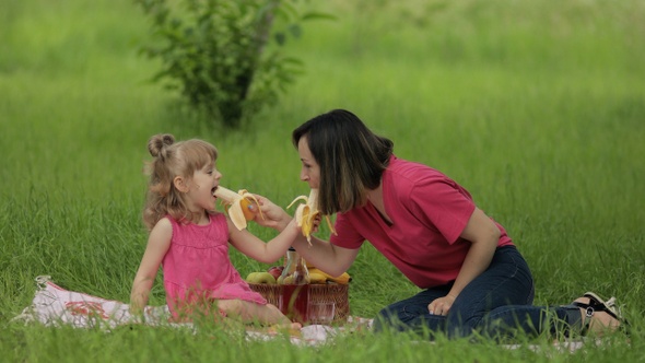 Family Weekend at Picnic. Daughter Child Girl with Mother on Grass Meadow Eating Bananas, Having Fun