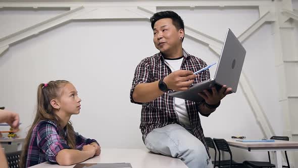 Korean Teacher Sitting on the Desk with Laptop in Hand Giving Lesson for Pupils