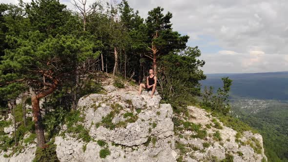 A Long-haired, Thin Man in a Black T-shirt and Shorts Sits on the Edge of a Cliff and Looks Out at