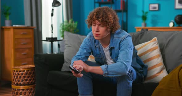 Teenager Boy Watching Television Using TV Remote While Sitting on Couch in Living Room at