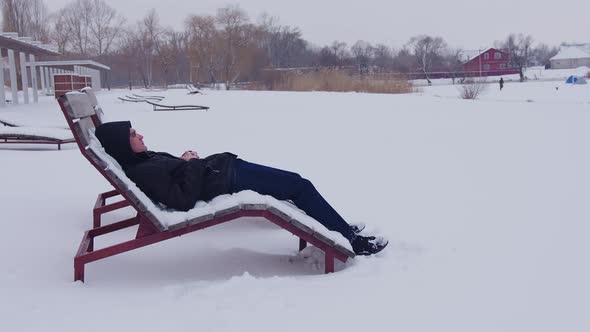 A Man Wearing Glasses Rests in a Sunbed in Winter Near a Frozen Pond in Winter in Snowy Weather and