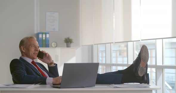 Businessman Making Phone Call Sitting with Legs on Table in Office