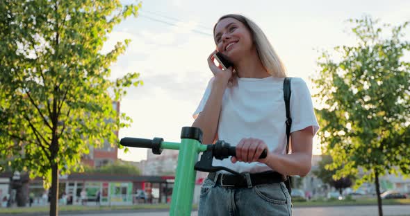 Happy woman stands with modern electric scooter talking on phone in city street