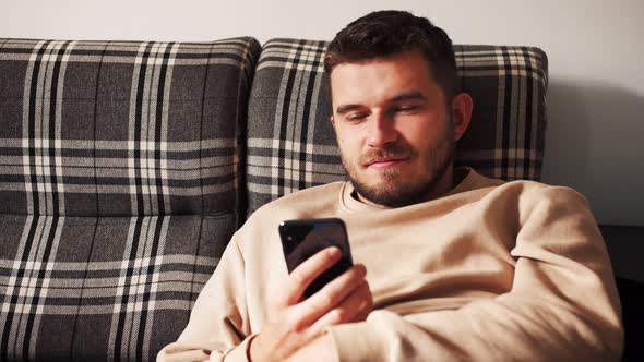 Gorgeous Caucasian Man Sitting on the Sofa Using Smartphone in the Modern Apartment Texting Message