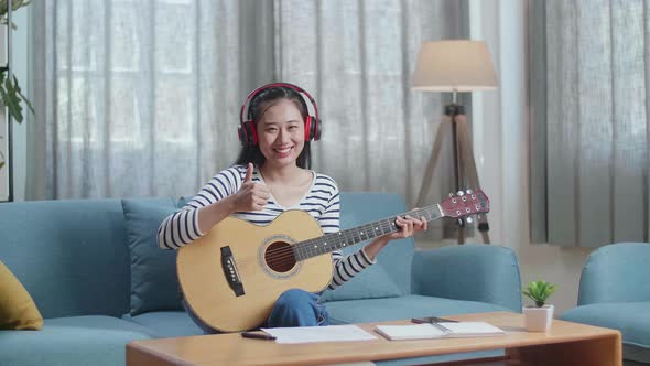 Asian Woman Composer With Headphones Smiling And Showing Thumbs Up To Camera While Playing Guitar