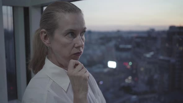 Close-up Portrait of Thoughtful Confident Woman Standing on Glazed Balcony at Dusk. Mid-adult