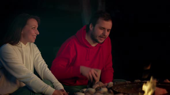 A Man Throws Firewood in a Fire. The Girl Sits By the Bonfire and Warms Hands