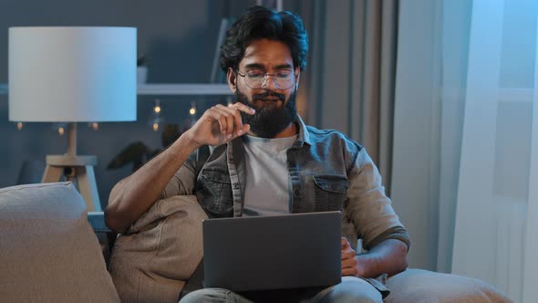 Adult Bearded Man in Glasses with Beard Sits in Evening in Dark at Home with Laptop Listening to