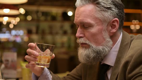 Close-up of a gray-haired bearded man enjoying whiskey alone in a bar