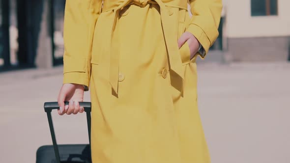 Beautiful Woman in a Yellow Raincoat Hat and Glasses Walks Down the Street with Luggage and Smiling