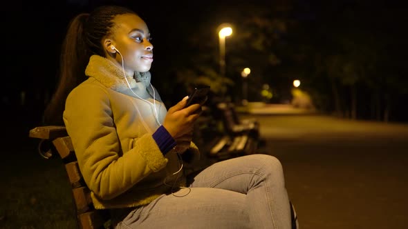 A Young Black Woman Listens to Music with Earphones on a Smartphone with a Smile in Park at Night
