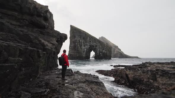 Man in Red Jacket By Rocks Overlooking Drangarnir Gated Arch in Faroe Islands