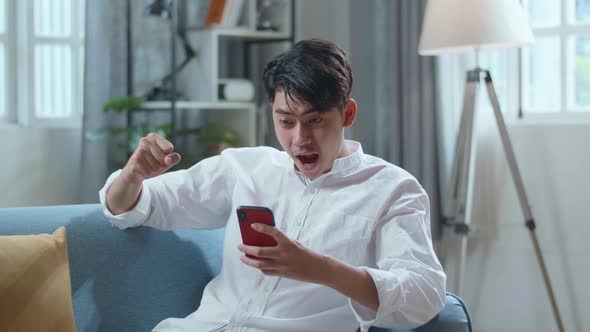 Happy Asian Man Celebrating While Lying On Sofa And Using Smartphone In The Living Room