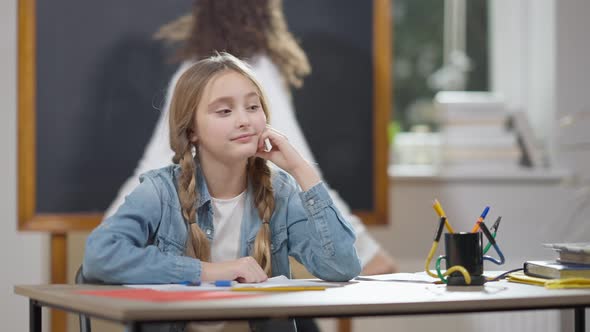 Bored Sad Caucasian Schoolgirl Sitting at Desk in Classroom with Blurred African American Classmate