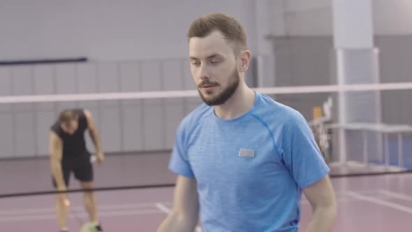 Young Smiling Man Showing Shuttlecock Serving at Camera with Blurred Caucasian Sportsman Stretching