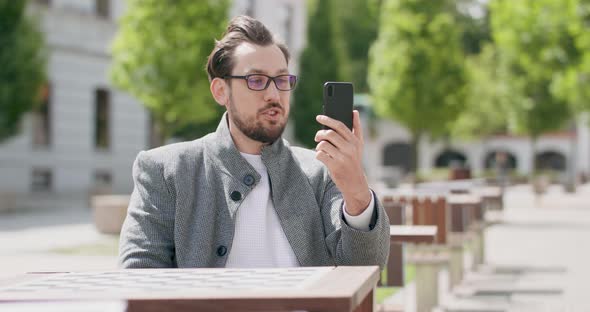 Young Man Wearing Glasses with Mustaches and a Beard is Sitting at the Table in the Square Chatting